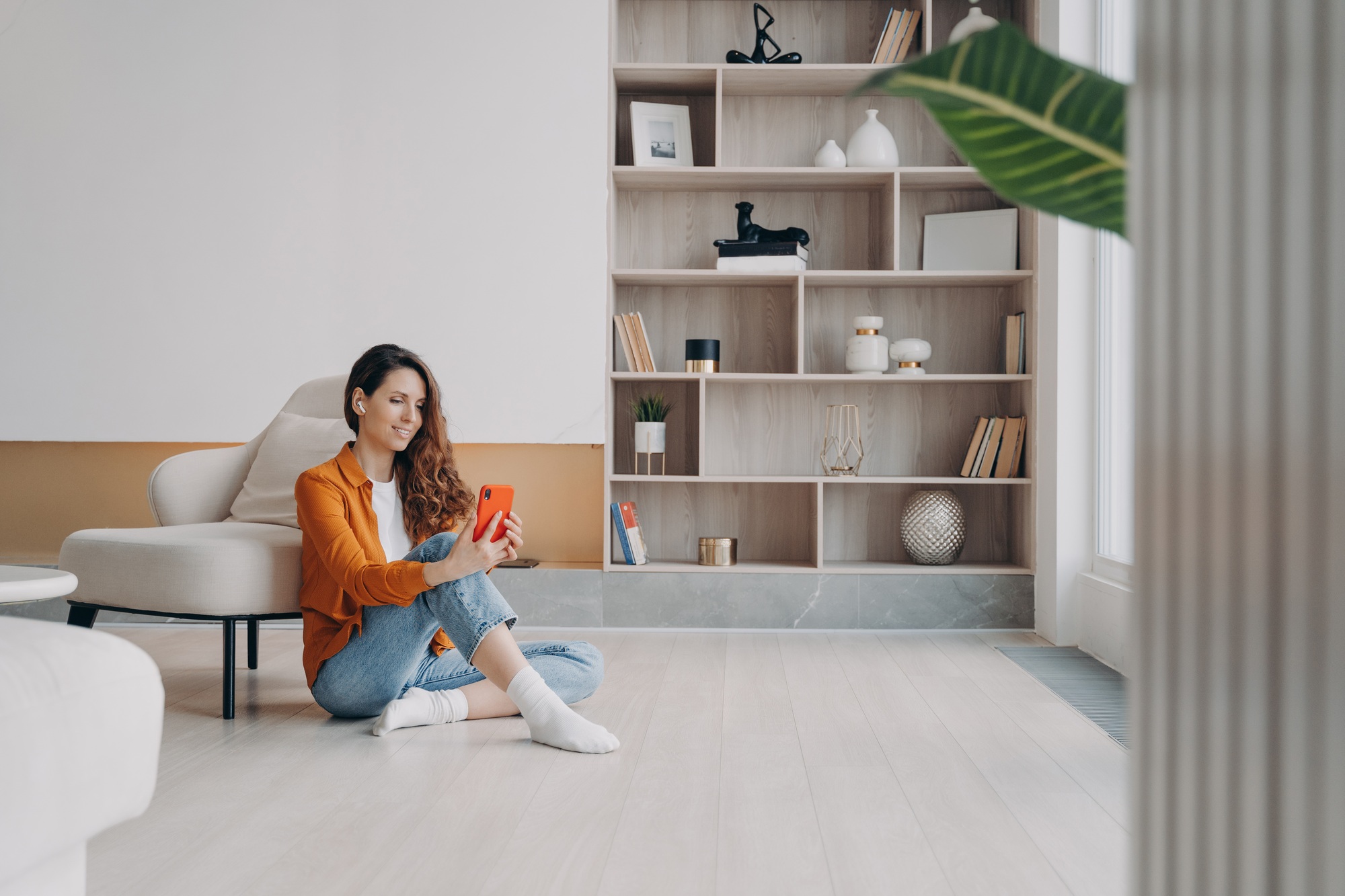 Happy woman homeowner sitting on heated floor, using smartphone apps in modern living room at home